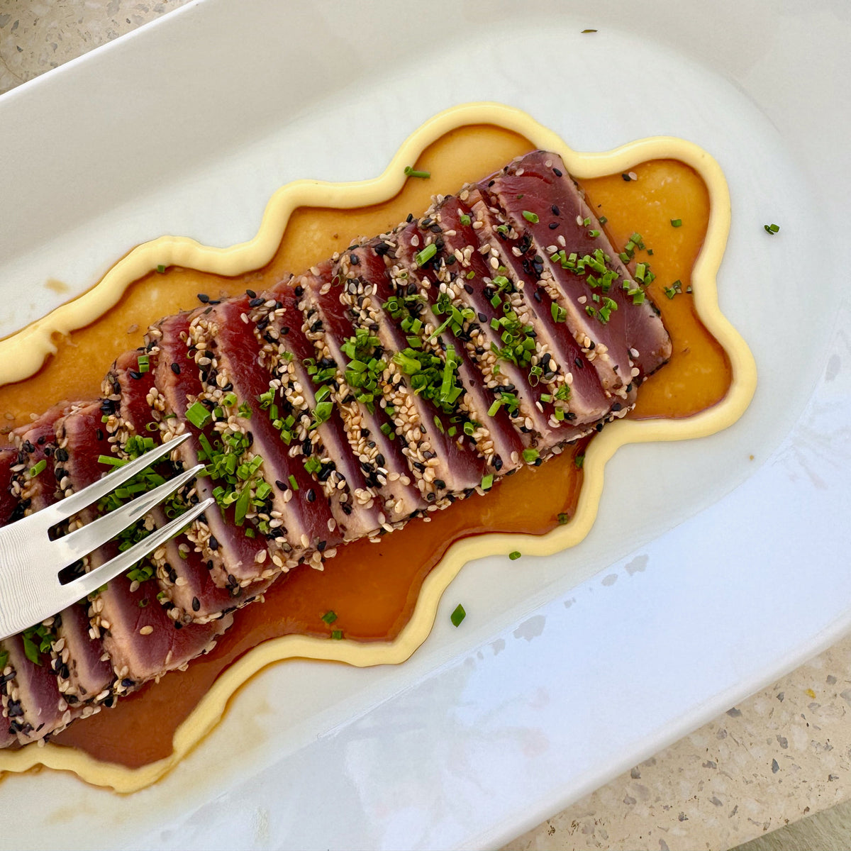 A close-up overhead shot of sliced raw tuna on a white plate, topped with sesame seeds, chopped green herbs, and a glossy sauce, with a fork entering from the left.