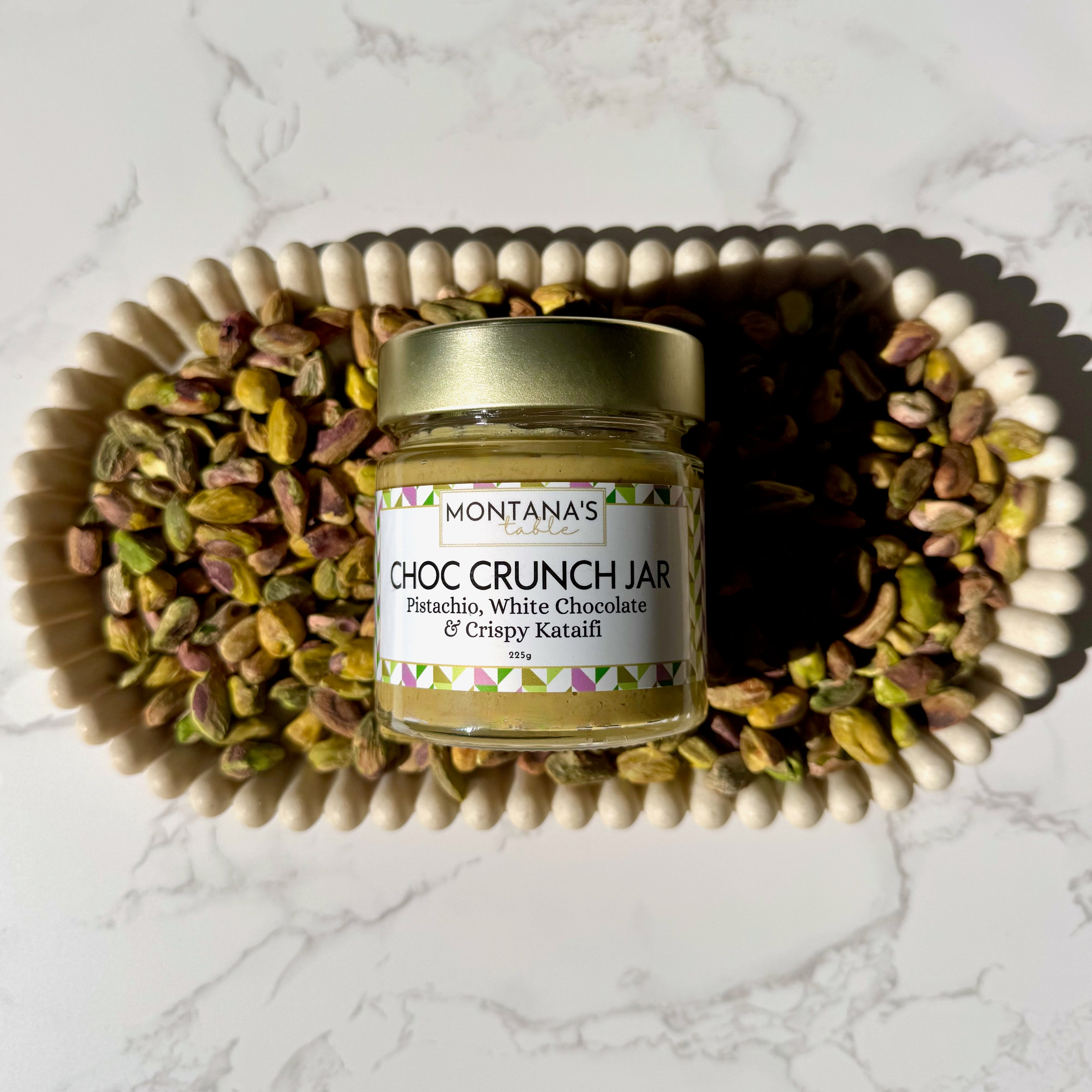 Jar of Montana's Table Choc Crunch Jar surrounded by pistachios on a marble surface